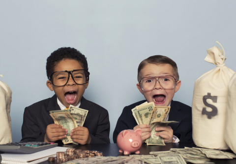 Two boys in suits and glasses shouting with excitement as they hold fistfuls of cash, beside a piggy bank and money bags