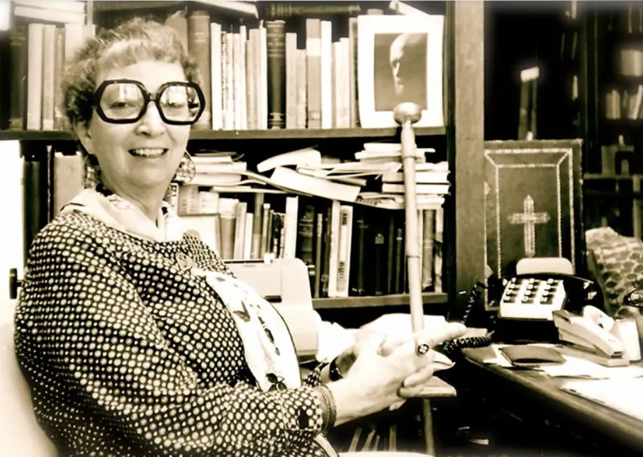 Madeleine L’Engle in large glasses at her desk, with bookshelves behind her
